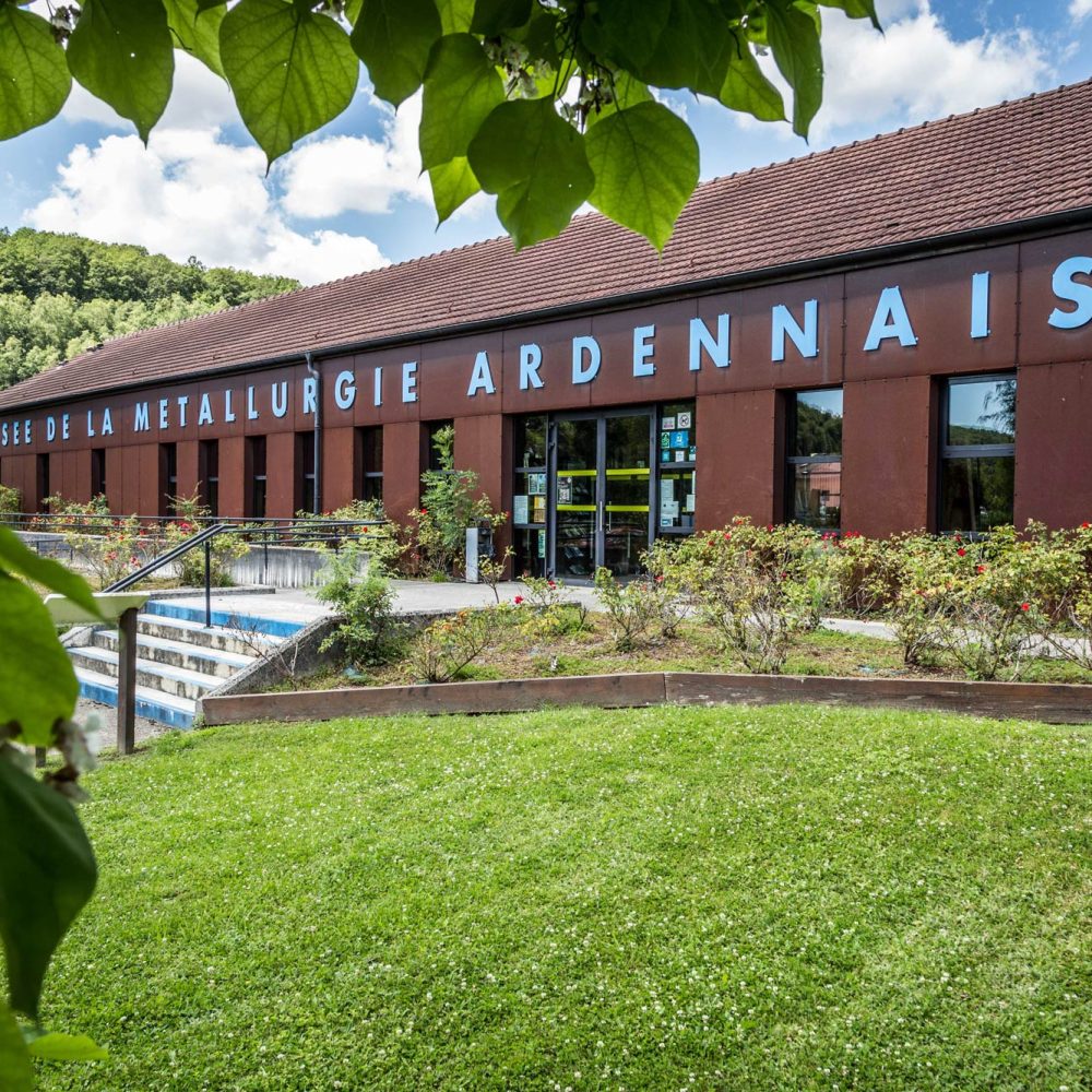 Façade du Musée de la Métallurgie Ardennaise avec son inscription en lettres bleues sur bâtiment en brique rouge, entouré de jardins paysagers dans le Parc naturel régional des Ardennes