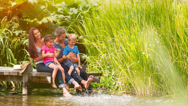 Famille d'habitants du Parc naturel régional des Ardennes assise sur un ponton en bois au bord d'un étang, parents et enfants profitant de la nature, végétation luxuriante