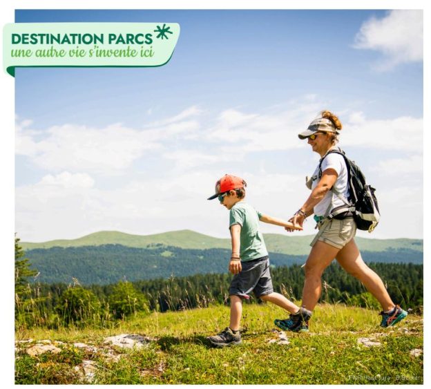Mère et enfant en randonnée main dans la main sur prairie alpine avec vue montagne avec la mention destination Parcs